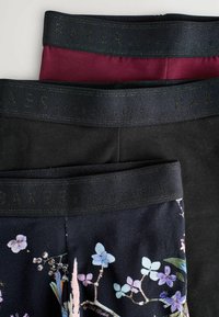 Tre paia di leggins impilati verticalmente. I colori includono bordeaux, nero e un tessuto nero con motivo floreale. Tutti hanno ampi elastici neri in vita.