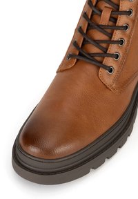 Hnedé kožené topánky s textúrovaným zvrškom, okrúhlou špičkou, kontrastnými čiernymi šnúrkami, kovovými očkami a hrubou čiernou gumovou podrážkou.