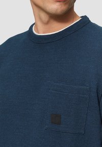 Marineblaue Struktur-Pullover mit Rundhalsausschnitt, ausgestattet mit einer Brusttasche und einem schwarzen Marken-Patch. Der Stoff hat ein gewebtes, geripptes Aussehen.