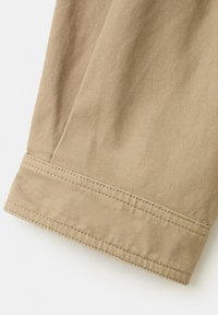 Gros plan sur une jambe de pantalon en tissu beige avec un ourlet à double couture et une texture de tissage diagonal visible.