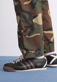 Sneakers in pelle nera con strisce marroni e dettagli bianchi, accompagnati da pantaloni con motivo camouflage. Suole in gomma e design minimalista.