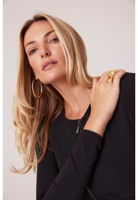 Czarna bluzka z długim rękawem i zamkiem przy dekolcie, w połączeniu z dużym złotym pierścionkiem i kolczykiem w kształcie kółka. Gładka, elegancka tekstura, bez widocznych wzorów.