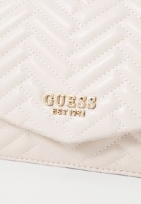 Quiltet beige læder clutch med guld "GUESS" logo og "EST 1981" detaljer. Har et herringbone mønster og en glat tekstur.