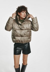 Beżowa, pikowana krótka kurtka z dużym kapturem, wyposażona w sznurki, zestawiona z czarnymi skórzanymi szortami i rękawiczkami.