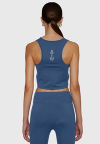Kvinna med brunt hår i en hästsvans som bär en blå ärmlös croptop med bokstäverna "ATW" och matchande högmidjade leggings, sedd bakifrån.