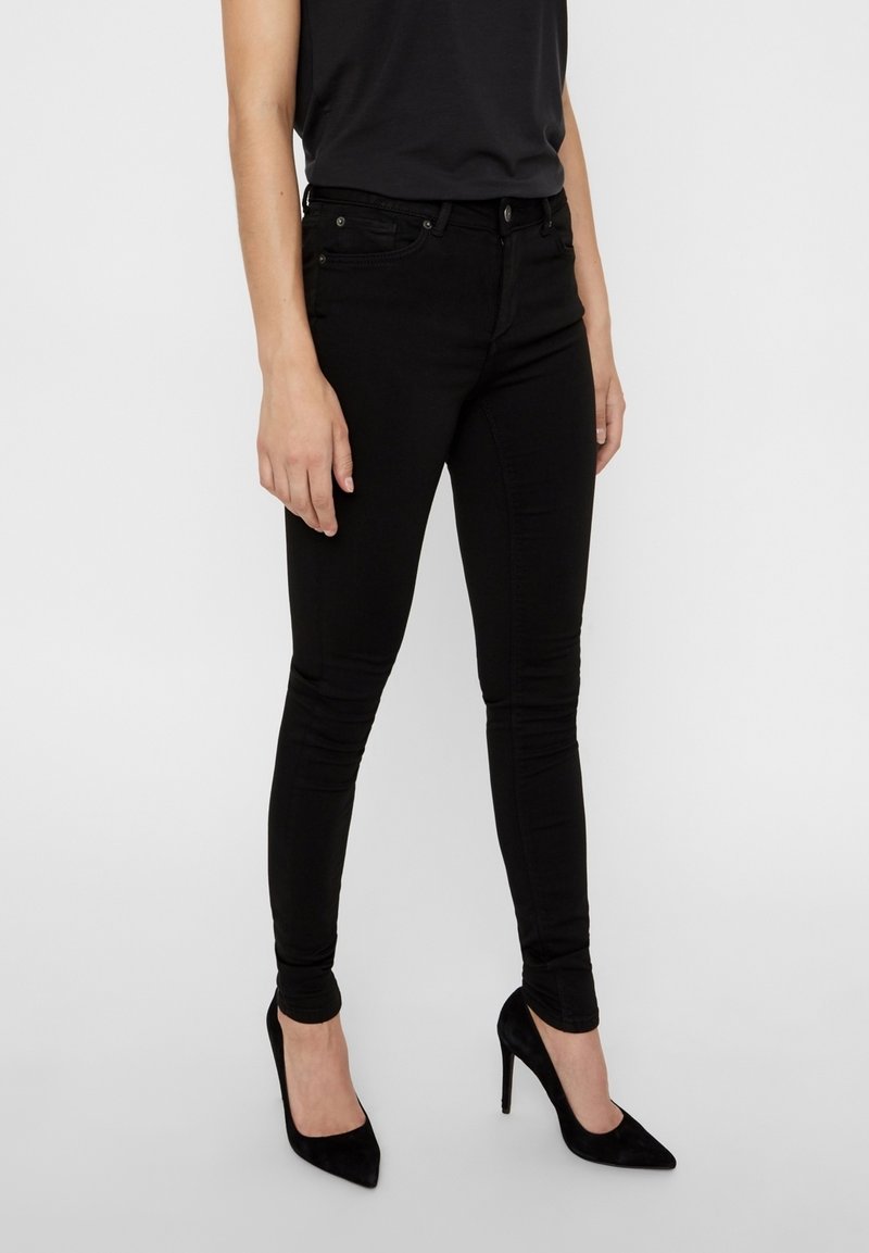 Jeans skinny negros con una textura de denim suave, que presentan cinco bolsillos, una cremallera y un corte ajustado, combinados con tacones altos negros.