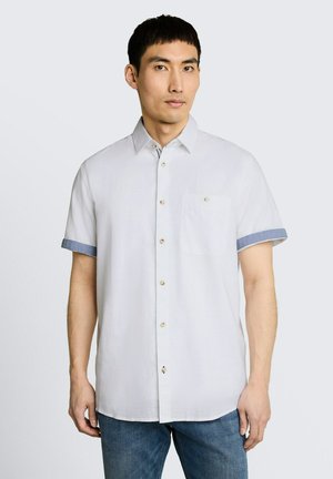 Mężczyzna ubrany w białą koszulę z krótkim rękawem z niebieskimi akcentami na mankietach oraz niebieskie jeansy, stojący na jednolitym jasnym tle.