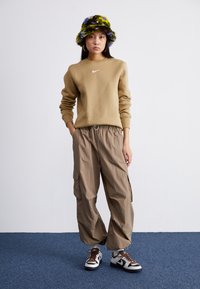 Sweatshirt bege com o logótipo branco da Nike, calças cargo bege com cós elástico e um chapéu de balde felpudo multicolorido. Ténis castanhos e brancos.