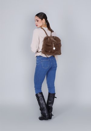 Vrouw met een bruine nepbontrugzak, een beige langemouwen-top, blauwe spijkerbroek en zwarte kniehoge laarzen, staand tegen een effen achtergrond.