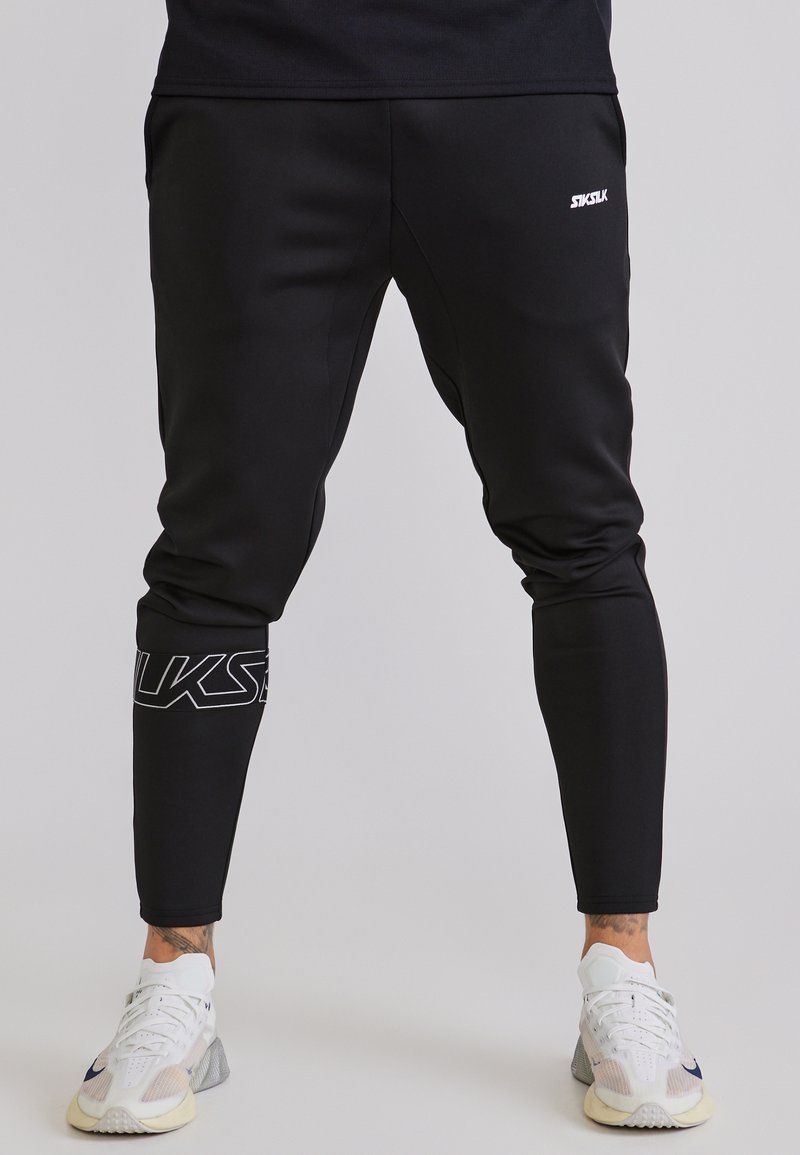Person wearing black tapered jogger pants with white logo on leg and ankle, paired with white athletic shoes, standing on gray floor.