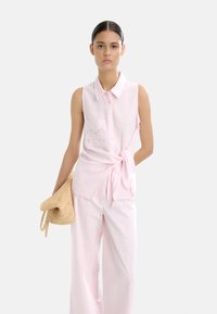 Jasnoróżowa bezrękawnikowa bluzka z kołnierzykiem, zapięciem na guziki i dekoracyjną teksturowaną kieszenią; zestawiona z jasnoróżowymi spodniami typu wide-leg i plecioną torbą.