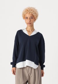 Tumes pullover V-kaelusega ja suuruse ülekaaluga, valge ja sinise triibulise kollase särgi kohal. Pehme, tekstuurne kangas. Silmapaistvad varrukad.