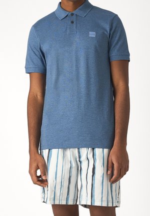 Blaues Poloshirt aus strukturiertem Stoff, mit einem Kragen und drei Knöpfen. Getragen mit gestreiften Shorts in hellblauen und dunkelblauen Tönen.