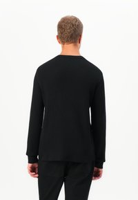 Homme avec des cheveux courts et bouclés portant un haut à manches longues noir et un pantalon noir, montré de dos sur un fond blanc.