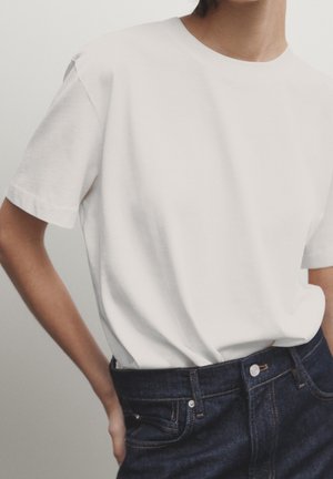 Person wearing a plain white short-sleeve t-shirt tucked into high-waisted dark blue denim jeans, with one hand resting on the hip.