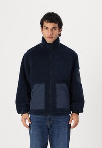 Marineblauer Fleece-Jacke mit hohem Kragen, Reißverschluss und zwei großen, kontrastierenden Stofftaschen auf der Vorderseite. Strukturiertes, gemütliches Material.