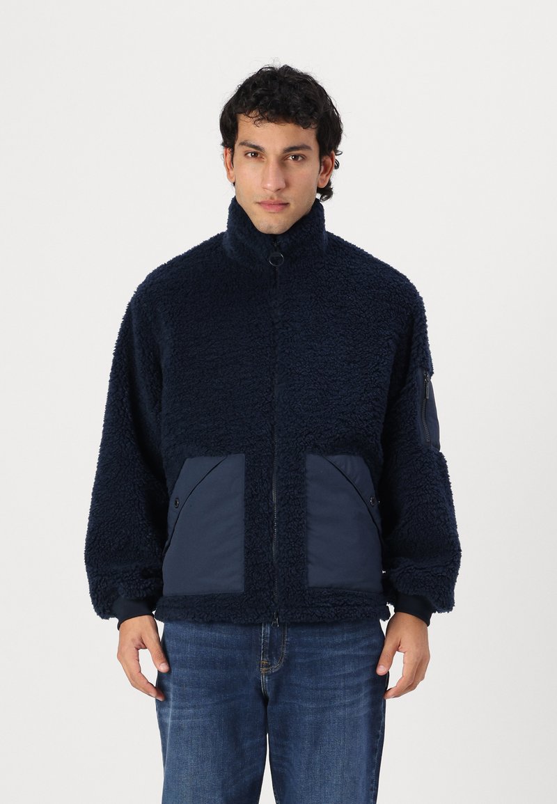 Marineblauer Fleece-Jacke mit hohem Kragen, Reißverschluss und zwei gro�ßen, kontrastierenden Stofftaschen auf der Vorderseite. Strukturiertes, gemütliches Material.
