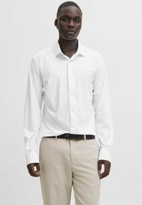 Camicia bianca a maniche lunghe con colletto appuntito, chiusura con bottoni e maniche con polsini. Abbinata a pantaloni beige chiaro e una cintura scura.