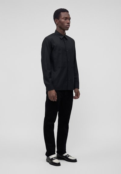 Homme debout portant une chemise noire boutonnée, un pantalon noir et des chaussures habillées noires et blanches devant un fond gris uni.