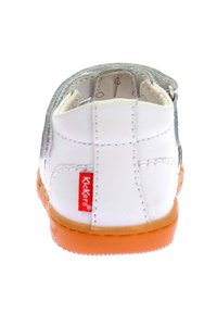 Vit barnskor med en slät lädertextur, silveraccenter, en orange gummisula och en röd "Kickers"-logotyp på sidan.