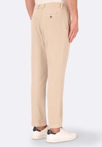 Pantaloni in misto cotone beige con una forma svasata, tasche posteriori con bottoni e un'etichetta piccola vicino alla vita. Indossati con sneakers bianche.