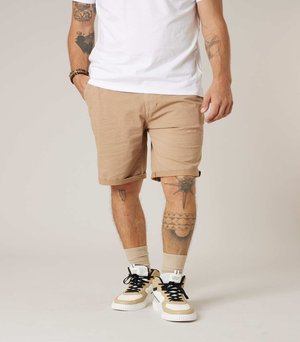 Man draagt beige korte broek met patroon, wit t-shirt, beige sokken en beige-witte sneakers met zwarte veters, toont tatoeages op armen en benen.