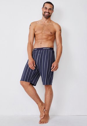 Lächelnder, oberkörperfreier Mann, barfuß stehend, trägt marineblaue, gestreifte Jockey-Shorts kniehoch vor einem schlichten weißen Hintergrund.