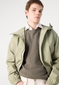 Light green waterproof jacket with a zip closure, featuring an adjustable hood and elastic cuffs, over a ribbed dark green sweater.