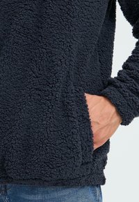 Pullover in pile navy con un aspetto morbido e tessurizzato. Presenta una tasca frontale e polsini a costine. Il materiale morbido offre calore e comfort.
