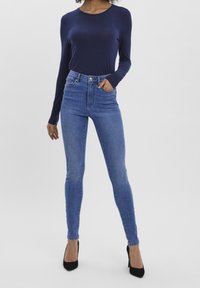 Jeans de mezclilla azul de cintura alta con una silueta ajustada, bolsillos frontales, presillas para cinturón, y combinados con una camisa de manga larga navy y tacones negros.