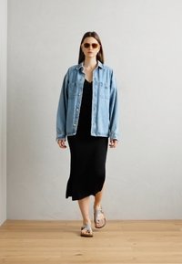 Denim ülepikkusega jakk, must ribiga kleit, prillid ja hõbedased sandaalid. Mudel kõnnib puidust põrandal lihtsa tausta taustal.
