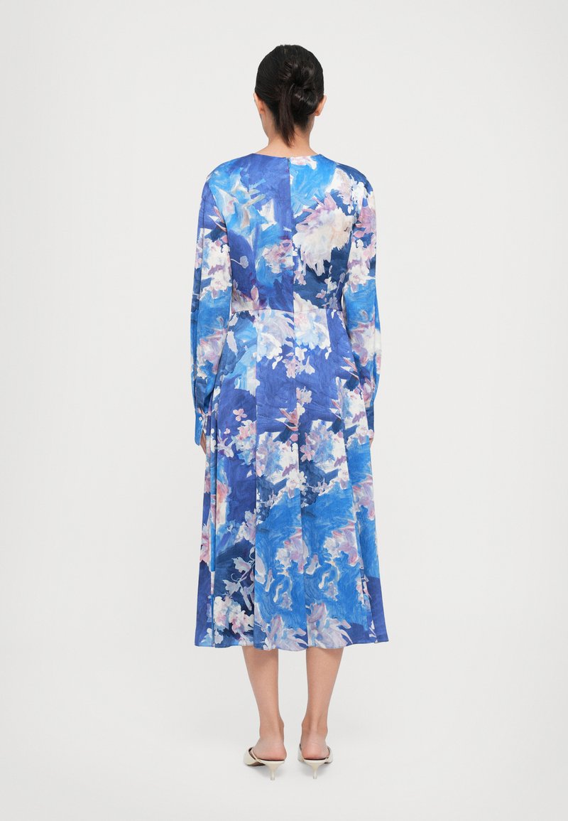 Vrouw met een knot in haar haar, gekleed in een langemouwen jurk tot halverwege de kuit met bloemen in blauw, wit en roze tinten, staand met haar rug naar voren gericht.