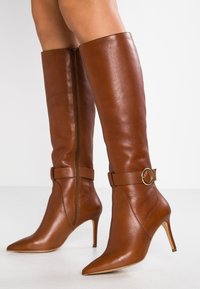 Bottes hautes en cuir marron jusqu'aux genoux, avec des bouts pointus, des talons fins, une sangle décorative avec un anneau doré, et une fermeture éclair latérale.