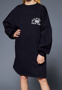 Námořnické šaty se širokými dlouhými rukávy, žebrovanými manžetami a bílým nápisem "HILFIGER 1985 NEW YORK" na přední straně. Měkký materiál.