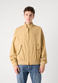 Beige bomberjakke med høj krave, lynlås lukning og to forlommer. Har elastiske manchetter og talje for et tætsiddende look.