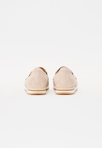 Beige semskede sko med lav hæl og hvid mellemsål med brune accenter. Designet er enkelt med glatte overflader og afrundede kanter.