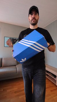 Blue Adidas shoebox with white three-stripe design. Box has a slightly worn edge, held in a person's hands, in a room with wooden flooring.