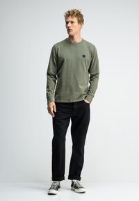 Langärmliges, olivgrünes T-Shirt mit dezenten Streifen und einem kleinen Logopatch. Kombiniert mit schwarzen Jeans und schwarz-weißen Sneakers.