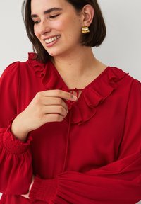 Rode blouse met gerimpelde halslijn en geplooide mouwen. Zachte stof, met een strikdetail bij de hals en elastische manchetten. Gouden oorbellen zichtbaar.