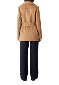 Kameelwollen jas met een riem in de taille, voorzien van een kraag en een gladde textuur. Draagt over donkere spijkerbroek. Achteraanzicht zichtbaar.