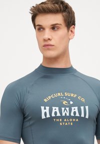 Kortermet surfeskjorte i mørk teal med en myk tekstur, med et frontgrafisk design i hvit og oransje som sier "Hawaii" og "Ripcurl Surf Co."