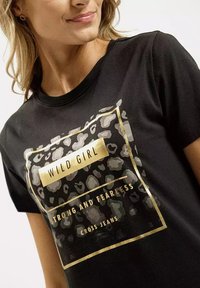 Czarny t-shirt z nadrukiem w złoto-białe cętki lamparta, z napisem "WILD GIRL" oraz tekstem "STRONG AND FEARLESS" umieszczonym wewnątrz prostokątnej ramki.