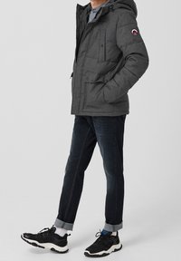 Giacca imbottita grigia con cappuccio, zip frontale, diverse tasche e patch con logo. Abbinata a jeans scuri e sneaker nere.