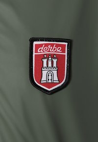 Patch logo Derbe rouge et blanc avec un emblème de château sur un fond en tissu vert foncé.