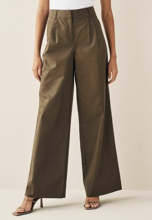 Vrouw draagt olijfgroene, wijde, hoge taille broek en bruine sandalen met bandjes en hoge hakken tegen een effen achtergrond.