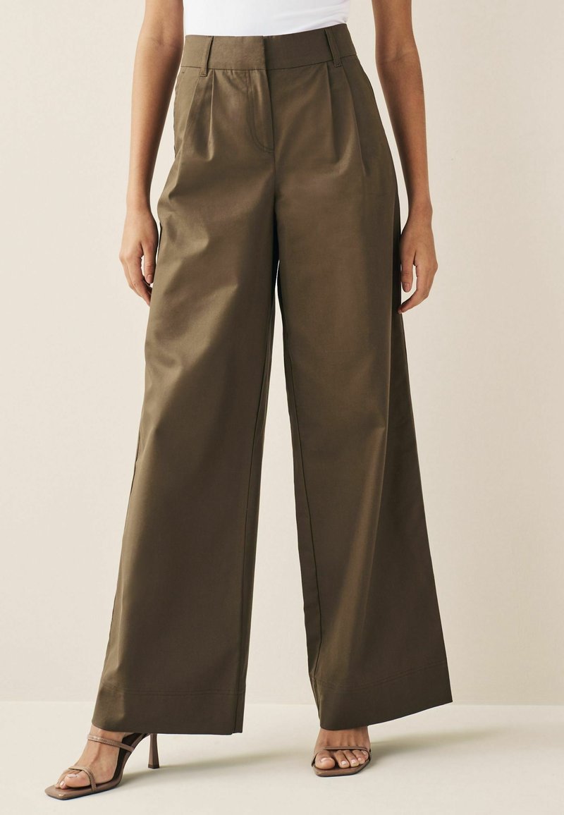 Femme portant un pantalon large taille haute vert olive et des sandales à talons hauts marron à brides sur un fond uni.