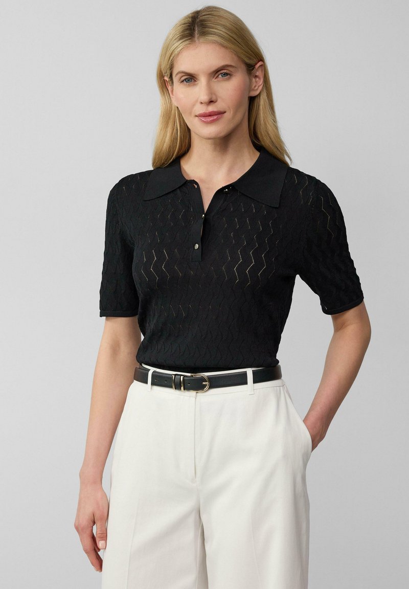 Zwart polo shirt met een gestructureerd zigzagpatroon, korte mouwen en een kraag. Gepaard met hoge witte broeken en een zwarte riem.