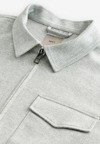 Lys grå zip-up jakke lavet af tekstureret stof. Har en struktureret krave og en frontlomme med en sort lynlås.