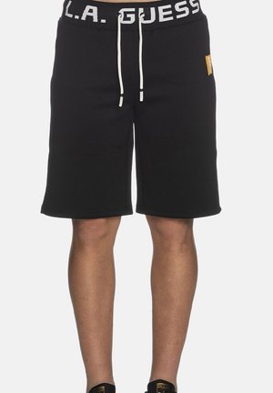 Pantaloncini neri fino al ginocchio con laccetto bianco e scritta "L.A. GUESS" sulla fascia della vita, indossati da una persona con gambe visibili e scarpe nere.