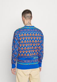 Felpa blu con intricati motivi floreali arancioni e dorati, polsini dettagliati e un bordo a contrasto all'orlo con il testo "CARLO COL".